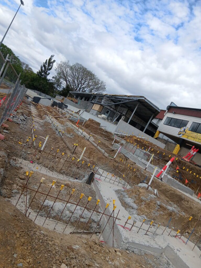 Excavated footings with reinforcing steel next to a grandstand under construction